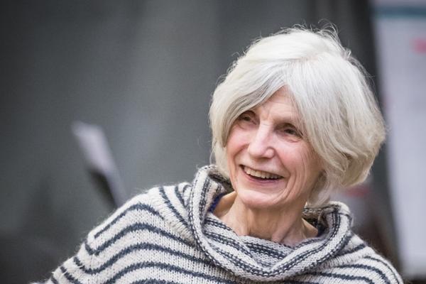 Caryl Churchill sits comfortably on a chair in a collaborative space, wearing a grey and blue striped jumper and holding a white mug with brightly coloured spots.